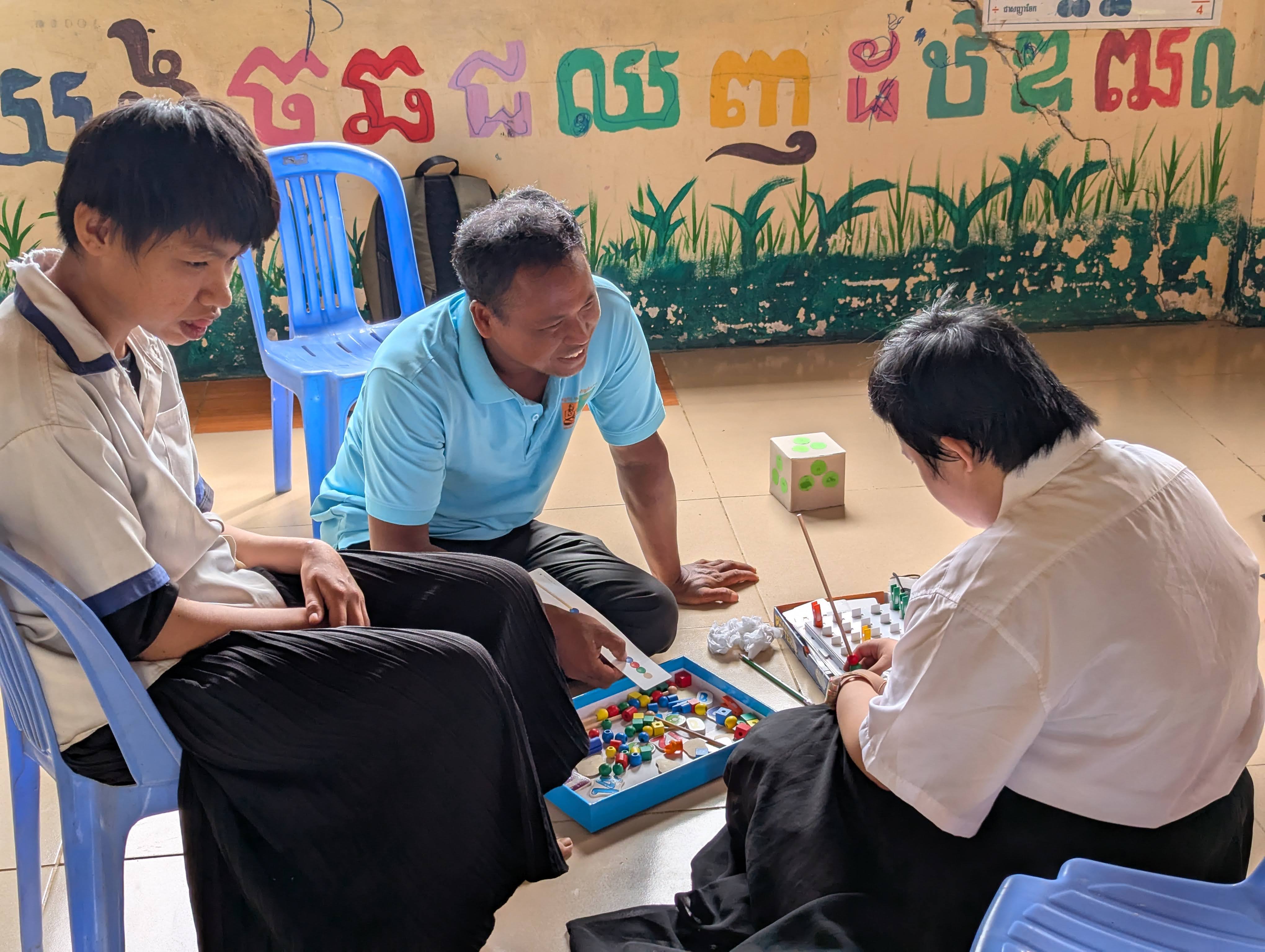 A teacher and two students sit on the floor, engaging in a board game with colorful pieces. They are surrounded by blue chairs, with a vibrant mural in the background.