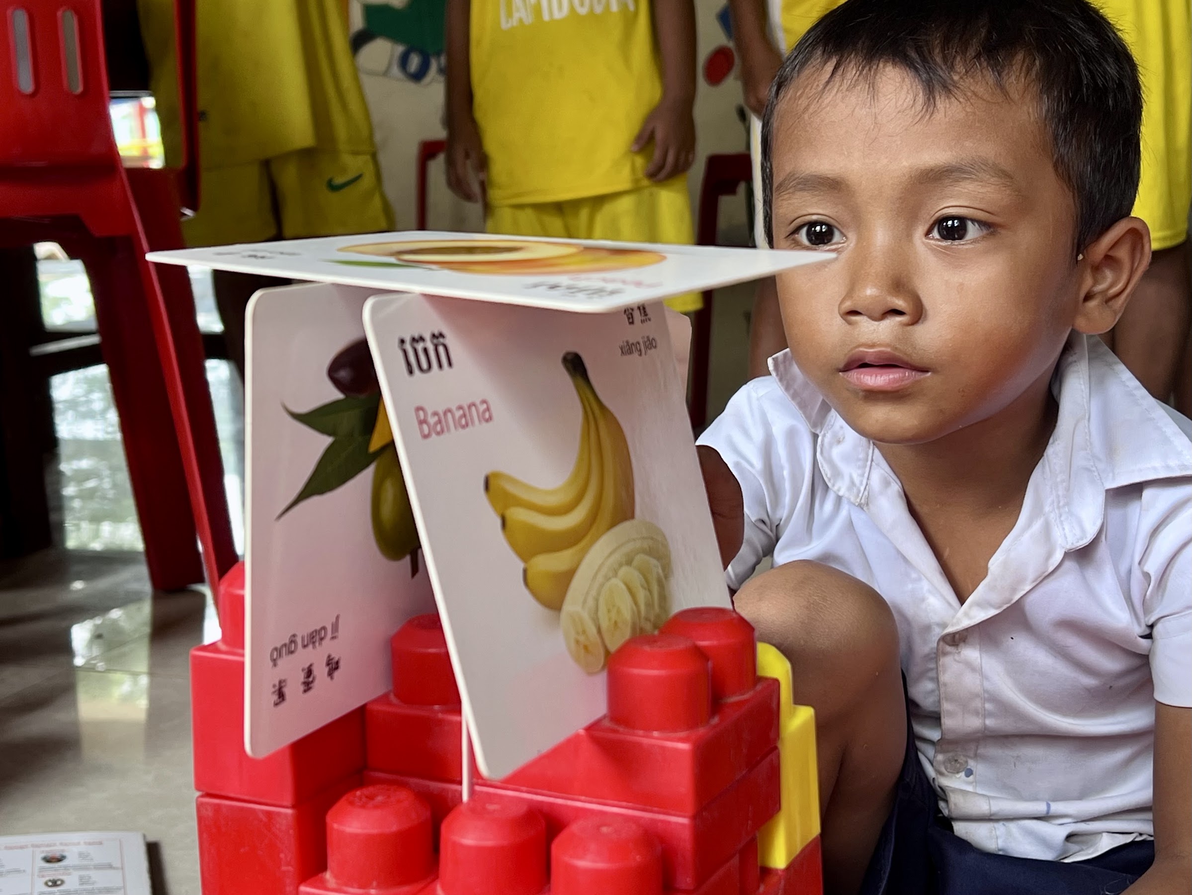 A young student builds three educational flashcards showing images of fruits and their names in English, Khmer, and Mandarin into a tower on top of red toy building bricks.