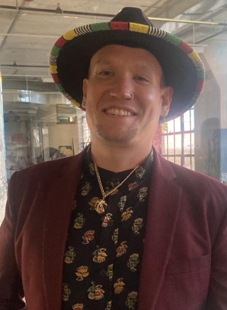 Daniel is wearing a black felt hat with bright colors on the edge. He has on a gold necklace, a black shirt with a bright pattern, and a maroon jacket. He is smiling.