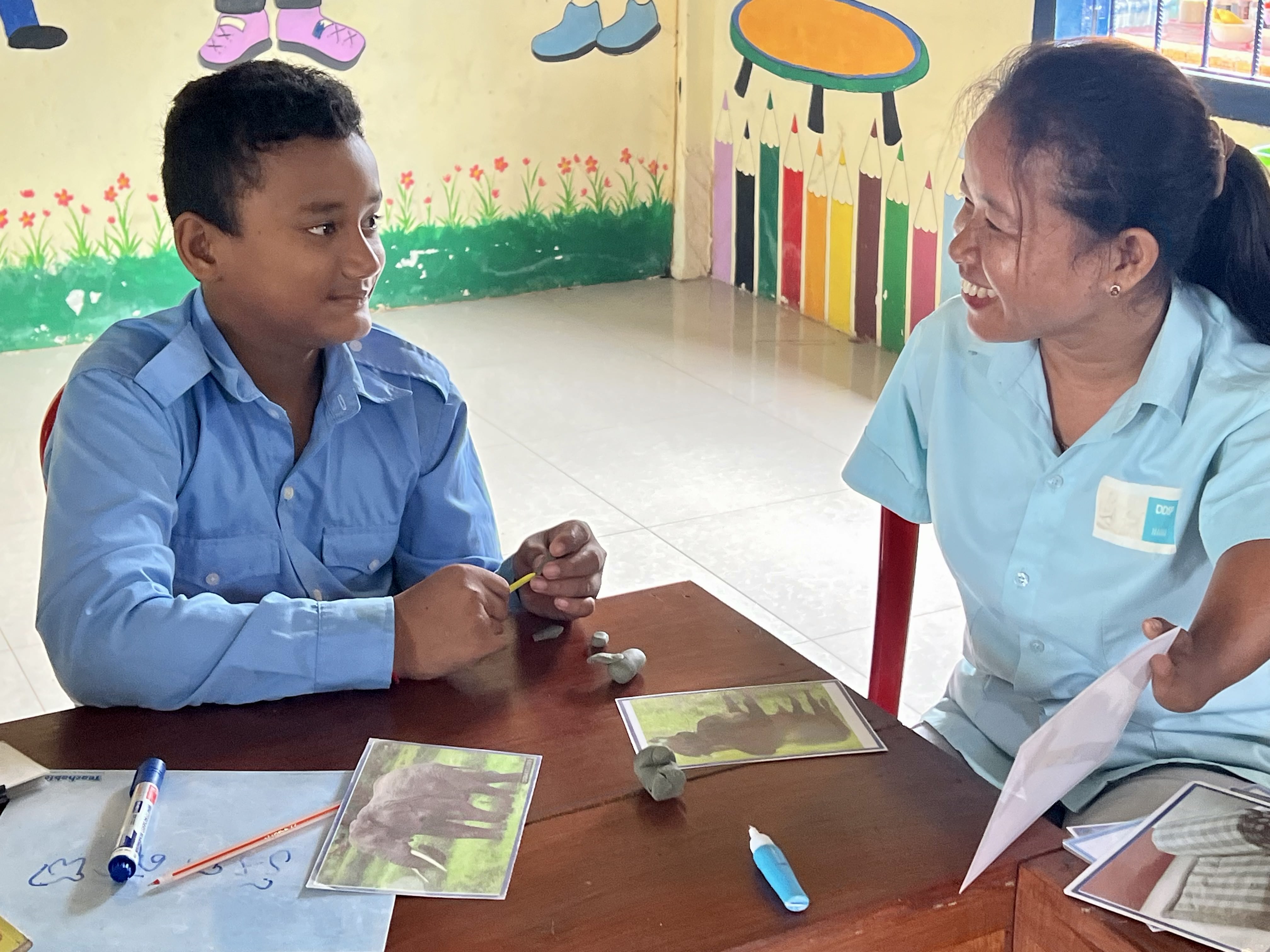 A young student and a teacher smile at each other. The teacher is showing the student an image of an elephant, and on the table there are small clay figurines resembling the animals.