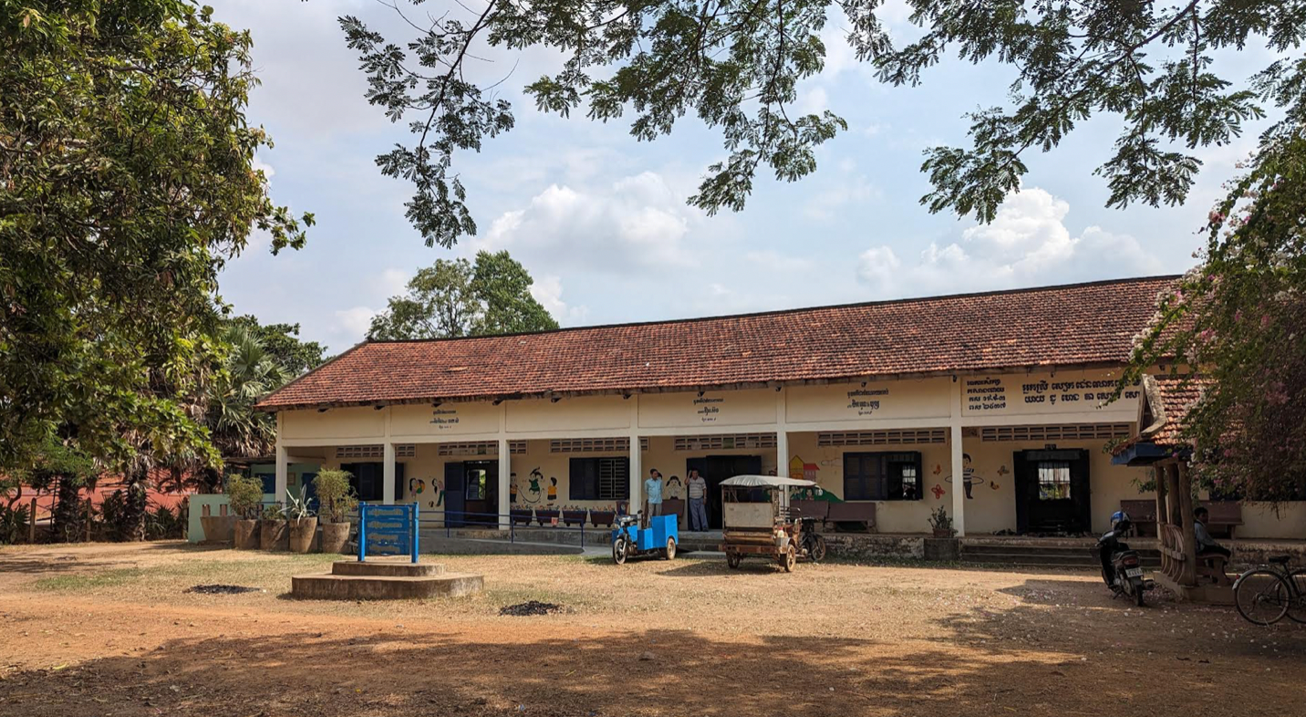 A single-story school building with a red-tiled roof and cream-colored walls in rural
            Cambodia. The building has a row of open windows and doors, with educational mural and
            Khmer script on the walls. An old tuk-tuk an small blue cart is parked in front. The yard is open
            with dry grass, shaded by large trees, and is sunny.