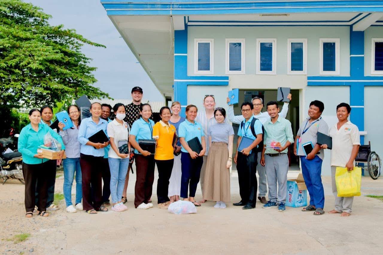 Daniel, Julie, Ashlyn, Hoa, Samnang, and thirteen other DDSP workers stand together in a group photo in front of the Inclusive Vocational Training Centre for People with Disabilities in Pursat, Cambodia. Several people are lifting up new tablet computers.